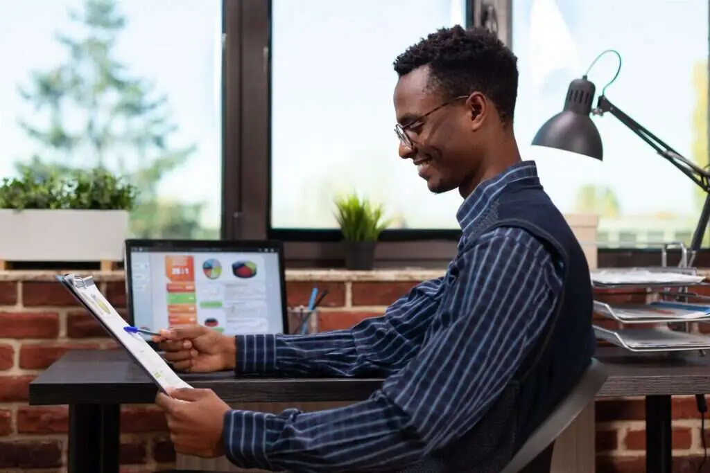 Payroll analyst reviewing a printed report against charts on a laptop screen
